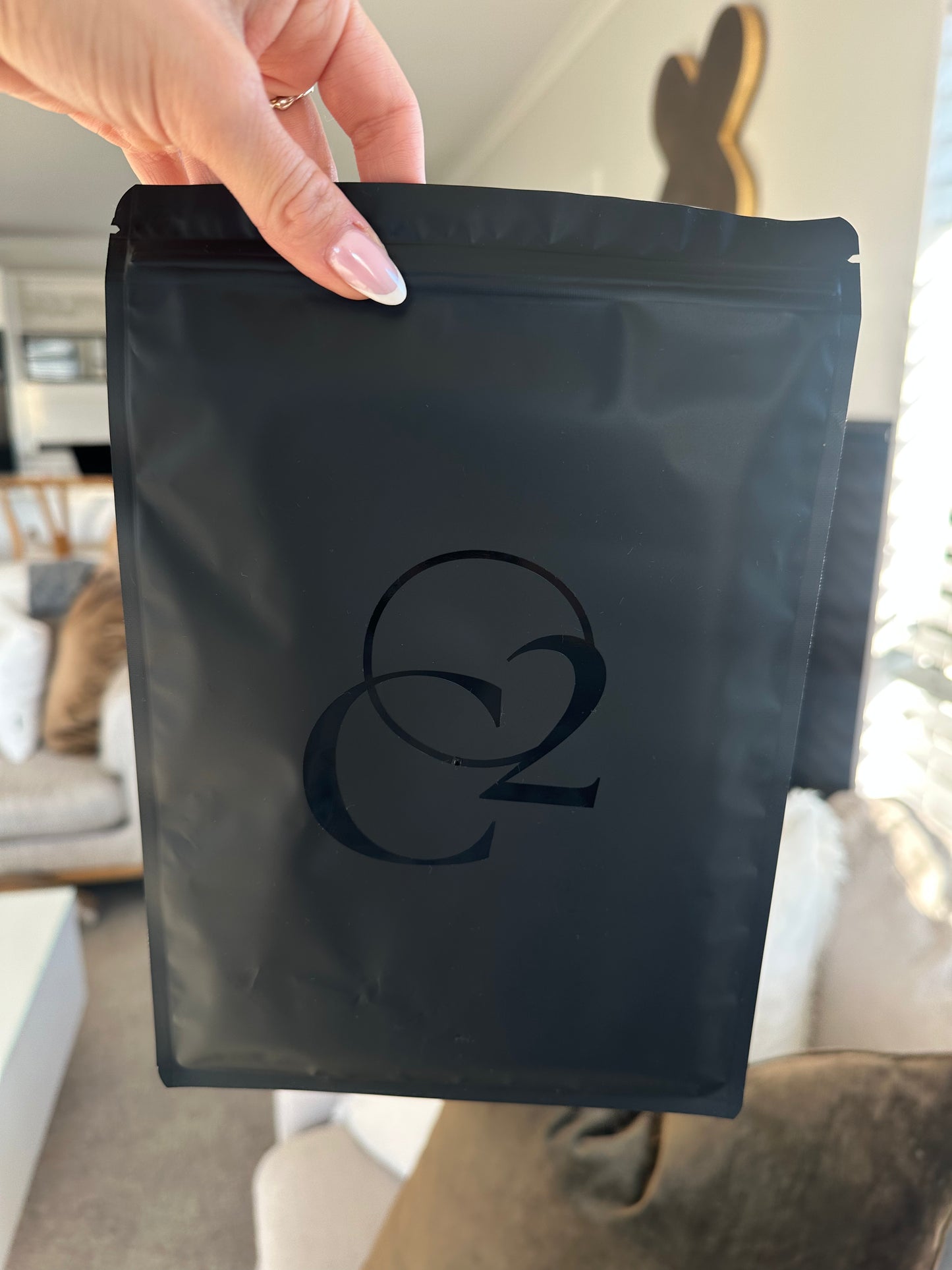A hand holds a matte black resealable stand-up pouch, filled with a carboxytherapy gel and face mask, with CO2 Society logo on the front, photographed indoors in a living room setting.